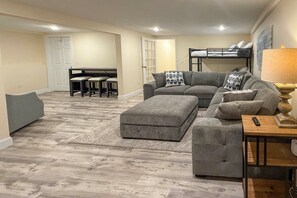 A modern living room with a grey sectional sofa, a grey ottoman, a wooden coffee table, and a black lamp. There is a dining area with a small table and chairs, and a bunk bed in the background. - Minutes Away From the Best Beaches in Cape Cod! (South Dennis)