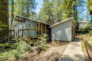 A rustic cabin with a wooden deck and a stone chimney, surrounded by dense forest. - Walk to Beach! Home w/ Sunroom in Anchor Bay (Gualala)