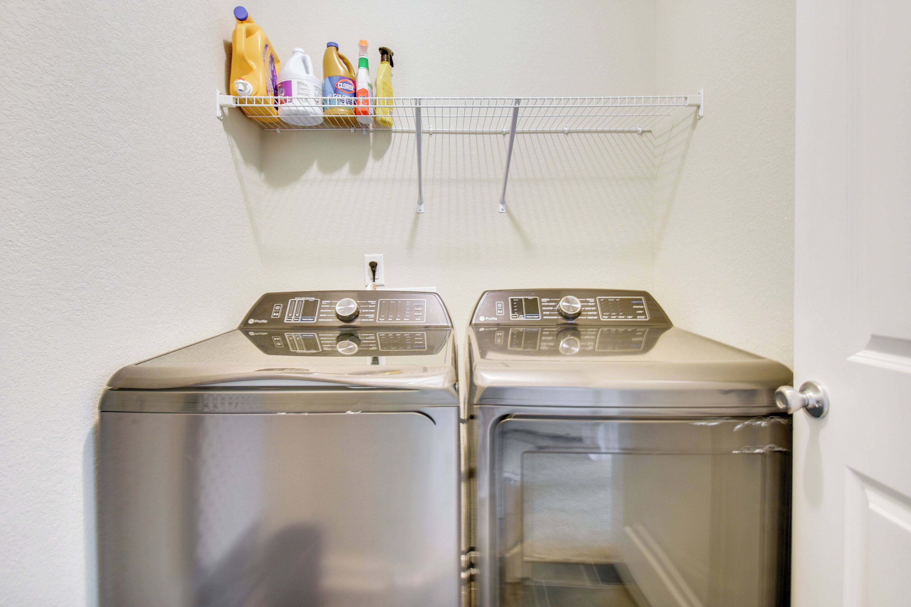 A laundry area with two stainless steel front-loading washing machines and a metal shelf above holding cleaning supplies.