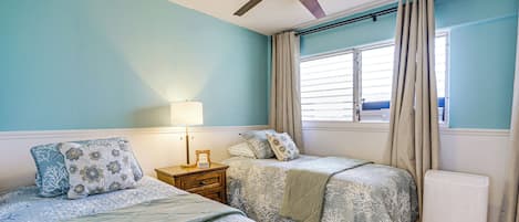 Un dormitorio con dos camas, ventilador de techo, mesita de noche, lámpara, ventana con cortinas y radiador.