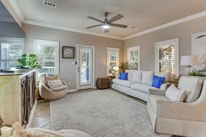Ein geräumiges Wohnzimmer mit Deckenventilator, großem Fenster mit Jalousien, einem weißen Sofa, einem beigefarbenen Sessel und einem gemusterten Teppich.
