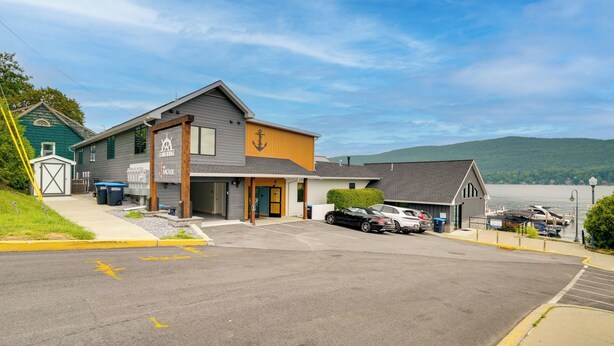 A two-story building with a sign for 'The Anchor' hotel. There is a parking lot with several cars, a grassy area, and a view of a body of water in the background. - Brand-New Luxury Lakefront Condo - 2200 Sq Ft! (Lake George)