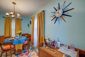 Een eethoek met een ronde tafel, stoelen, een kroonluchter, gordijnen en een wandklok.