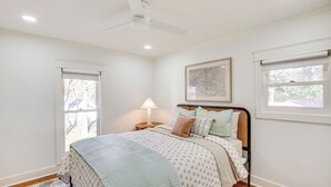 Ein Schlafzimmer mit einem Bett, einem Deckenventilator, einem Nachttisch mit Lampe und einem Fenster mit Blick auf Bäume.