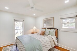 Ein Schlafzimmer mit einem Bett, einem Deckenventilator, einem Nachttisch mit Lampe und einem Fenster mit Blick auf Bäume.