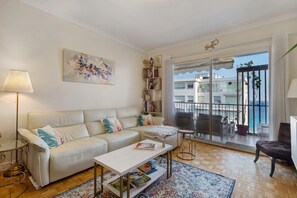 A modern living room with a sofa, coffee table, and a bookshelf. There is a large window with a view of buildings and a balcony. - Sea View 2 Bed & 2 Bath - Highly Rated, with Wi-Fi and Air Conditioning (Nice)