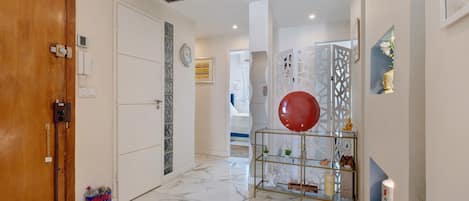 A hallway with a wooden door, a white wall with a clock, a glass shelf with decorative items, and a marble floor.