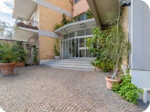 A courtyard with a paved walkway leading to a building entrance, surrounded by potted plants and a small tree. - Keys of the Vatican by A Picture of Rome (Roma)