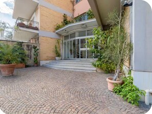 A building entrance with a brick pathway leading to a glass door, surrounded by potted plants. - Keys of the Vatican by A Picture of Rome (Roma)