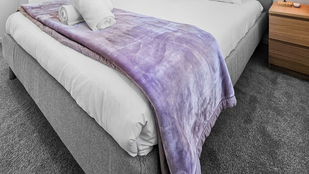 A modern bedroom with a grey bed, white bedding, a purple blanket, and a wooden nightstand with a lamp. - Waipuna Living | 3-Bed Townhouse | Spacious Home (Mount Wellington)