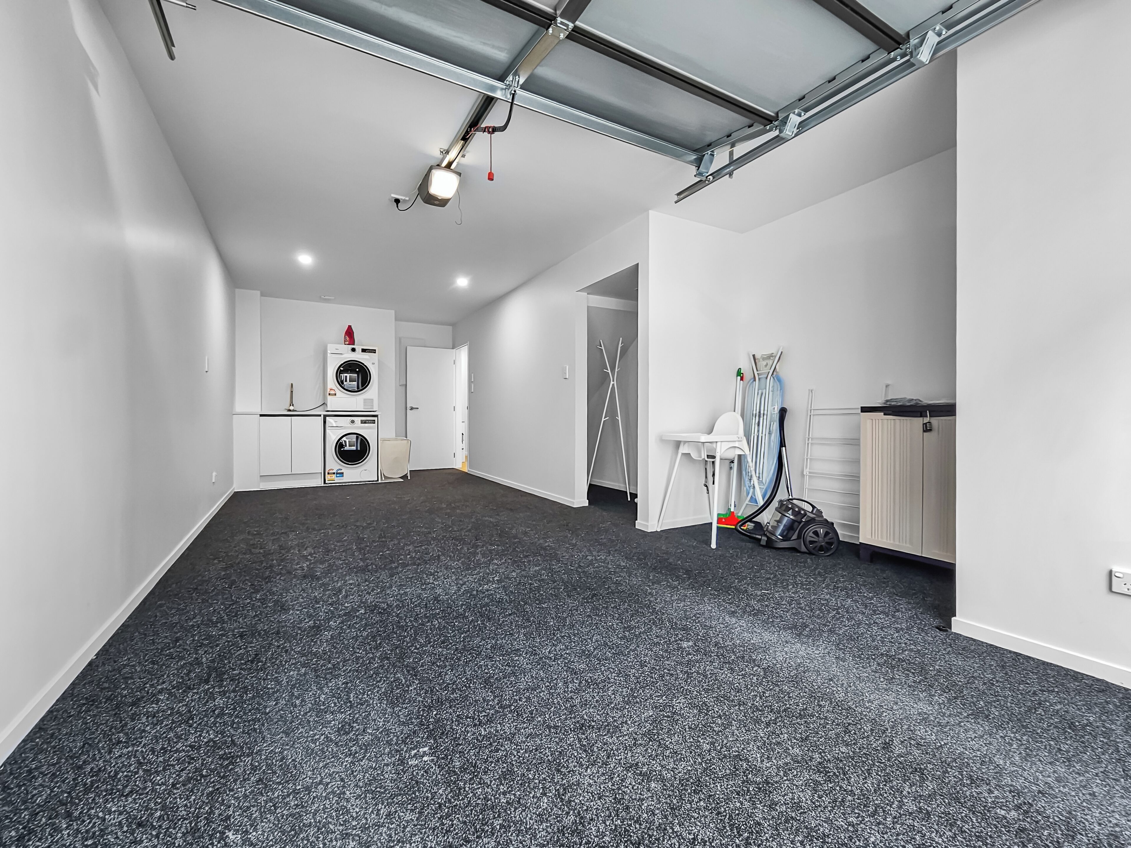 A spacious room with a dark carpet, a ceiling with exposed ductwork, a white door, and a laundry area with appliances.