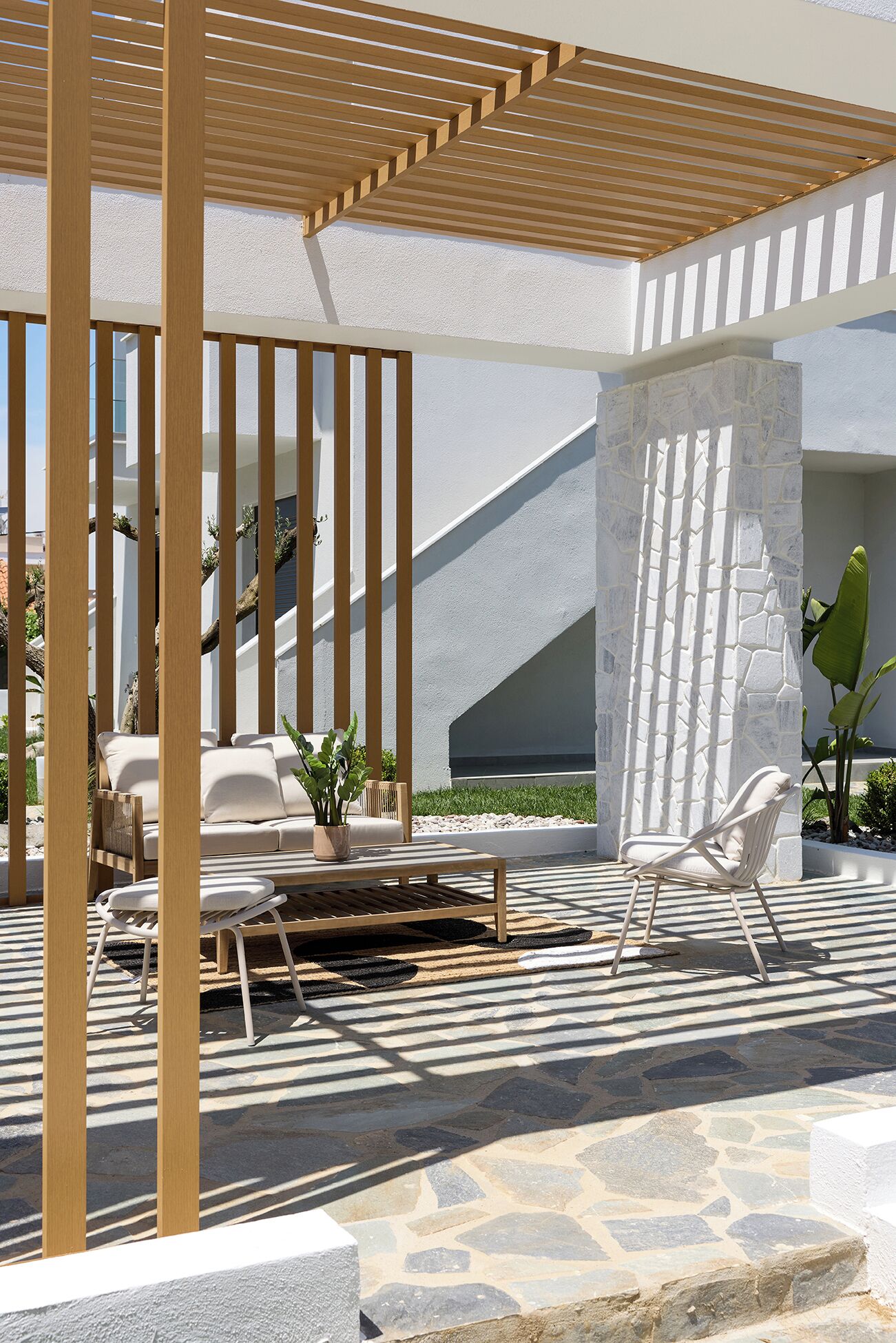 An outdoor seating area with a pergola, stone flooring, and a white wall with a geometric design.