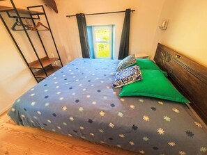 A bed with a patterned bedspread, two patterned pillows, and a view of a green outdoor area through a window. - Gîte 2 "Le Bateau Yves" by Interhome (Blainville-sur-Mer)