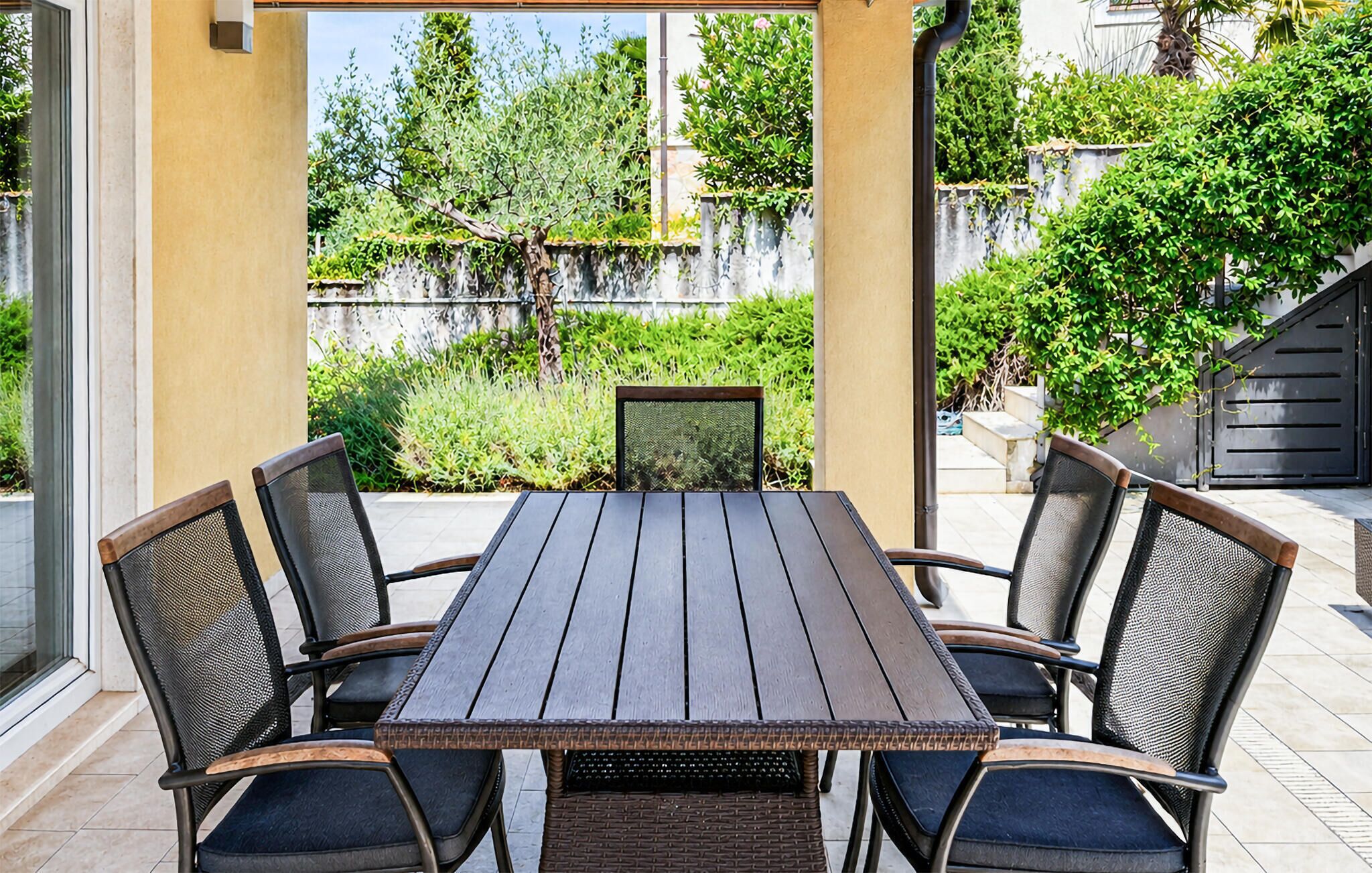 Une table rectangulaire pour l’extérieur, accompagnée de six chaises, entourée d’un mur de couleur beige et de verdure.