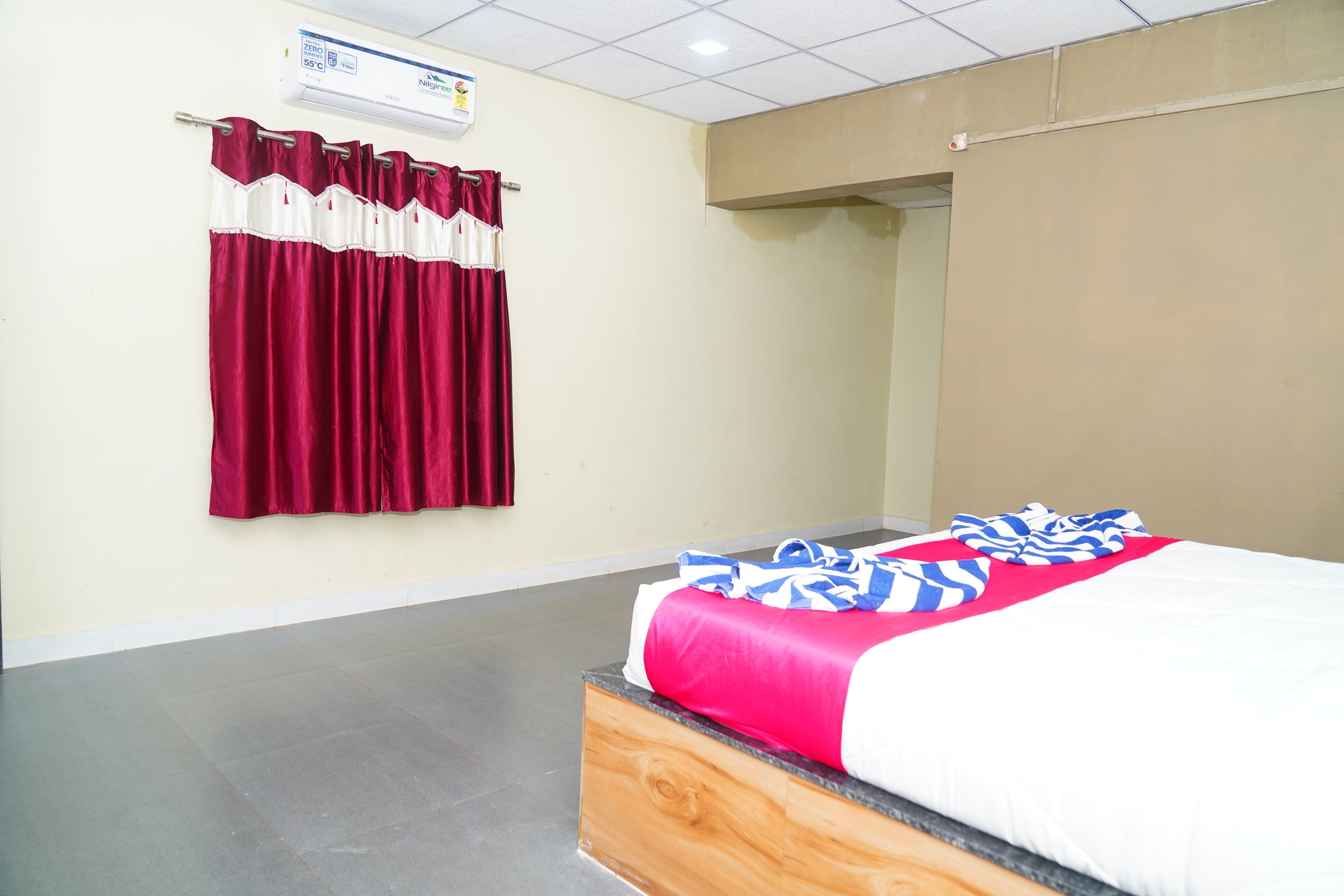 A hotel room with a bed, a window with curtains, a wall-mounted air conditioner, and a tiled floor.