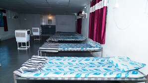 A row of single beds with patterned bedding, a standing heater, and a wall-mounted fan in a room with a concrete ceiling and red curtains. - Kesar Villa (Bhojde)