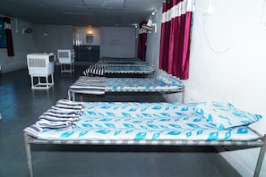 A row of single beds with patterned bedding, a standing heater, and a wall-mounted fan in a room with a concrete ceiling and red curtains.