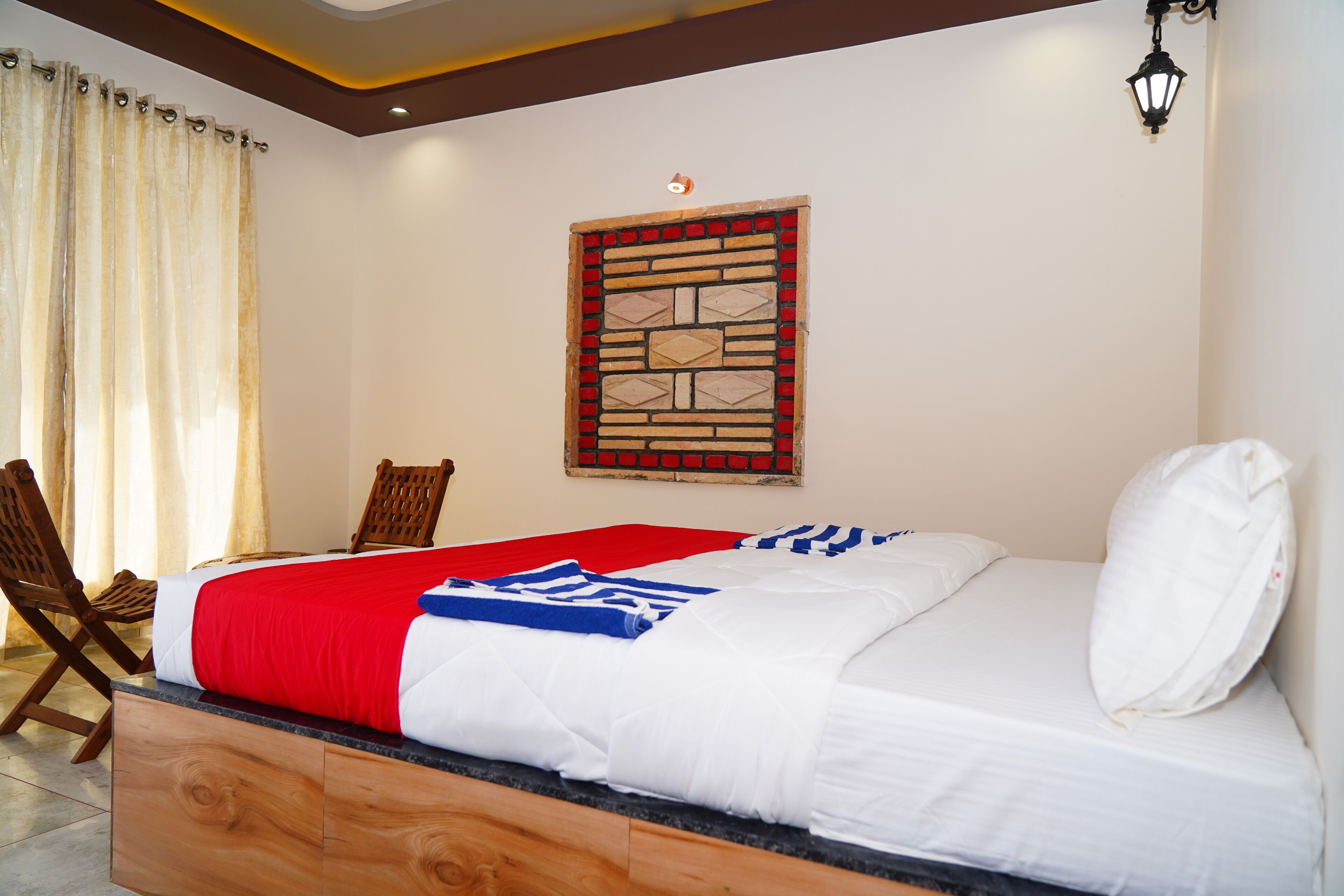 Habitación de hotel con cama, cabecera de madera, colcha blanca y roja, almohada blanca, silla de madera, ventana con marco decorativo y lámpara de pared.