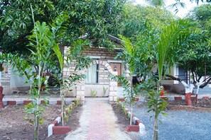 A pathway leading to a small building with a stone facade, surrounded by lush greenery and young plants. - Kesar Villa (Bhojde)