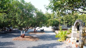 A paved driveway leading to a gated entrance with a stone archway, surrounded by lush greenery and trees. - Kesar Villa (Bhojde)