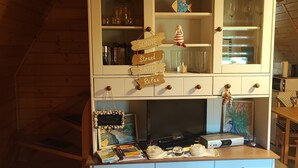 L’intérieur d’une cabane en bois comprend un coin cuisine, un téléviseur et une étagère où sont disposés des objets décoratifs.