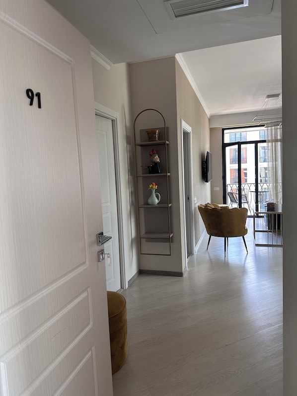 A hallway with a door labeled '91', a shelf with decorative items, a sofa, and a view of an outdoor balcony.