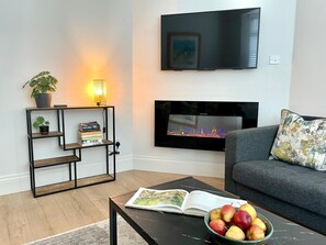 A living room with a fireplace, a flat-screen TV, a bookshelf, and a coffee table with a book and a bowl of fruit. - The Cottage - Centre of Newcastle Co.Down (Newcastle)
