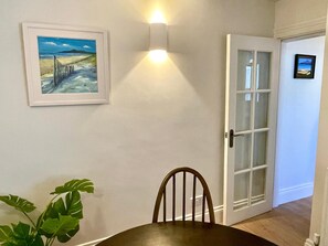 A dining area with a table, a chair, a framed painting, a wall-mounted light, and a door with glass panels. - The Cottage - Centre of Newcastle Co.Down (Newcastle)