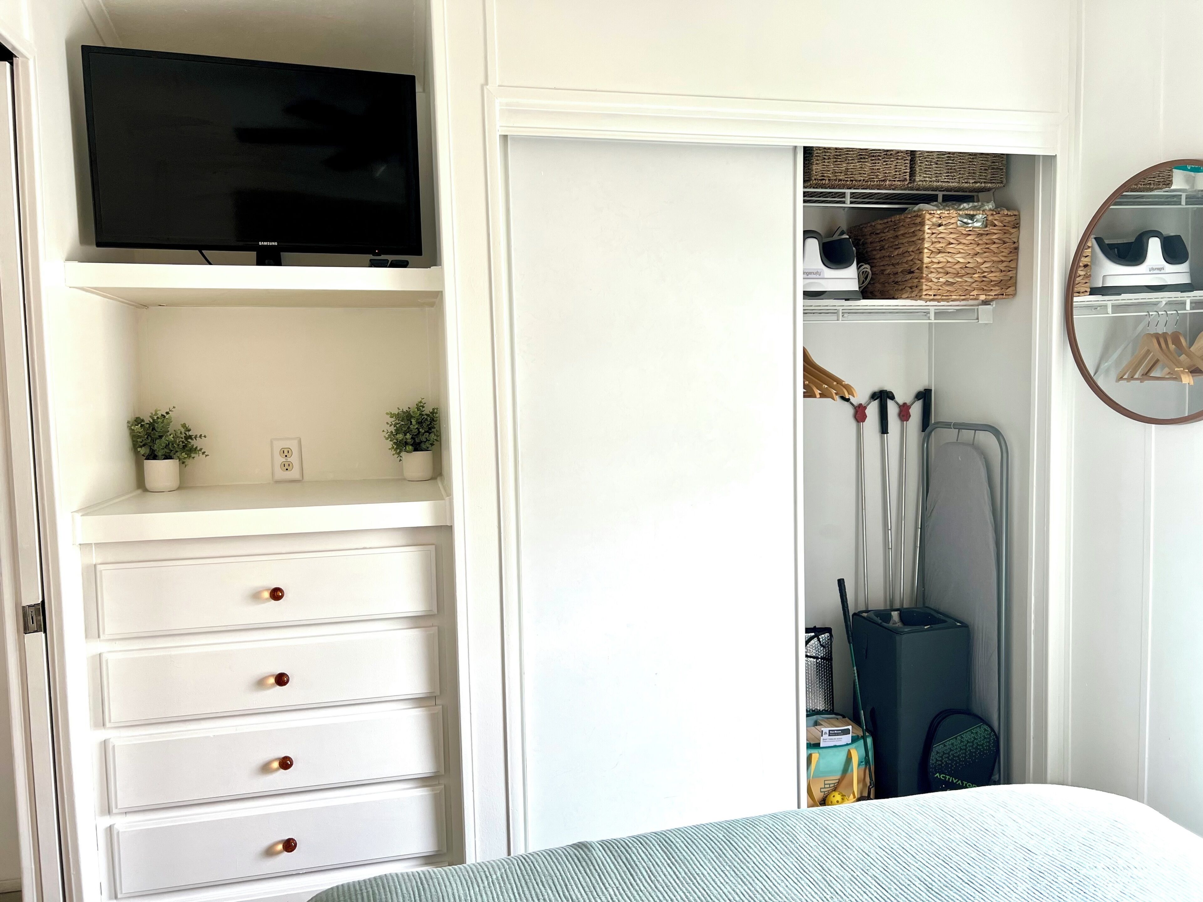 Une chambre à coucher avec un lit, un téléviseur posé sur une étagère, une armoire contenant des vêtements suspendus et une petite plante.
