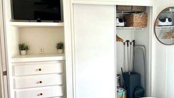 Une chambre à coucher avec un lit, un téléviseur posé sur une étagère, une armoire contenant des vêtements suspendus et une petite plante.