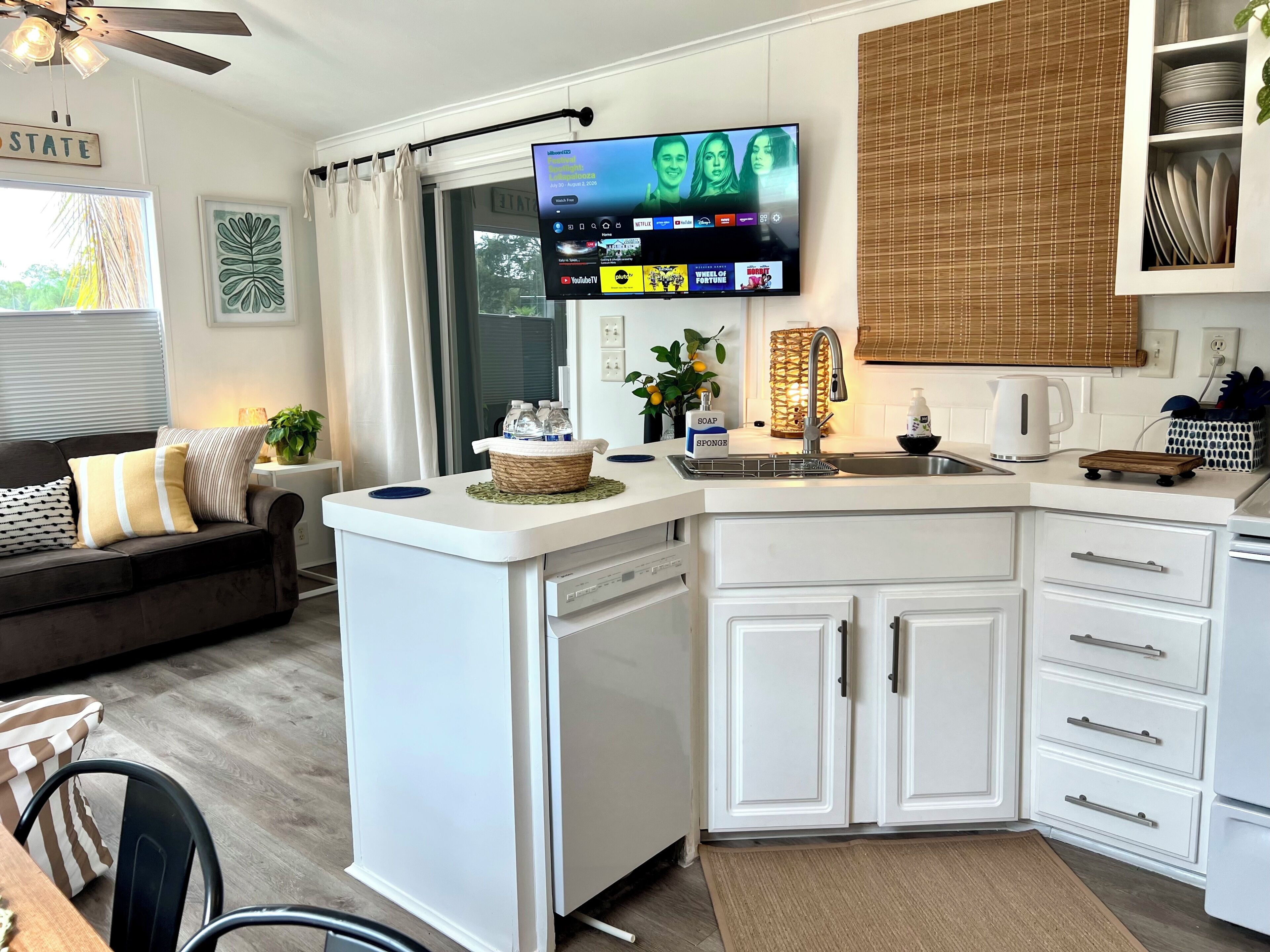A modern kitchen with a built-in dishwasher, a flat-screen TV mounted above the counter, and a dining area with a sofa and a coffee table.