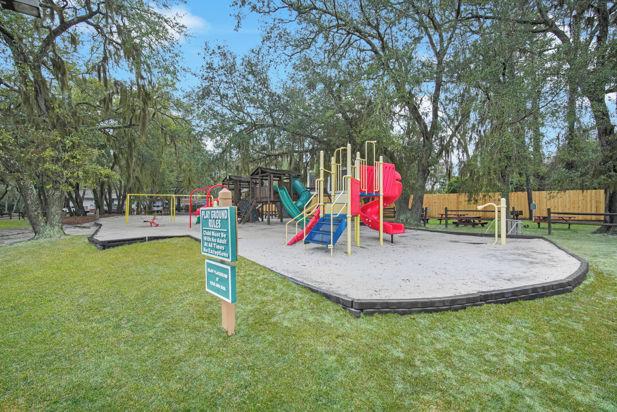 A playground with a slide and climbing structures, surrounded by trees and a wooden fence.