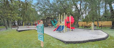 A playground with a slide and climbing structures, surrounded by trees and a wooden fence.