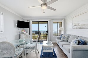 A room with a glass table, white wicker chairs, a grey sofa, a ceiling fan, and a TV mounted on the wall. - AR 308 Hilton Head Beach &Tennis Beach Lovers Only (Hilton Head Island)
