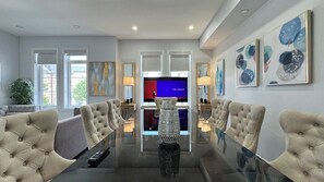 Una sala moderna con una mesa de comedor de cristal, sillas tapizadas en beige, un televisor de pantalla plana grande, arte abstracto en la pared y una planta en maceta.