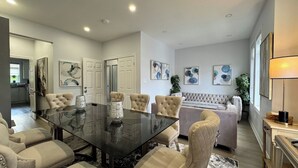 A modern living room with a glass-top dining table, beige upholstered chairs, a tufted sofa, and abstract wall art.