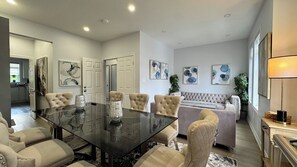 Una sala de estar moderna con una mesa de comedor de vidrio, sillas tapizadas en beige, un sofá capitonado y arte abstracto en la pared.