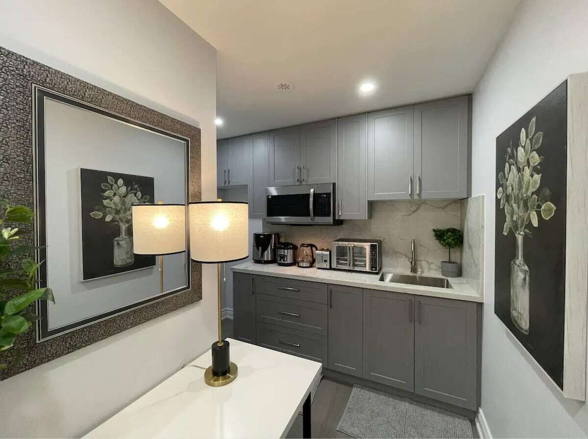 A compact kitchen with grey cabinets, a microwave, a toaster, and a sink. There are two wall-mounted lamps on a white table.