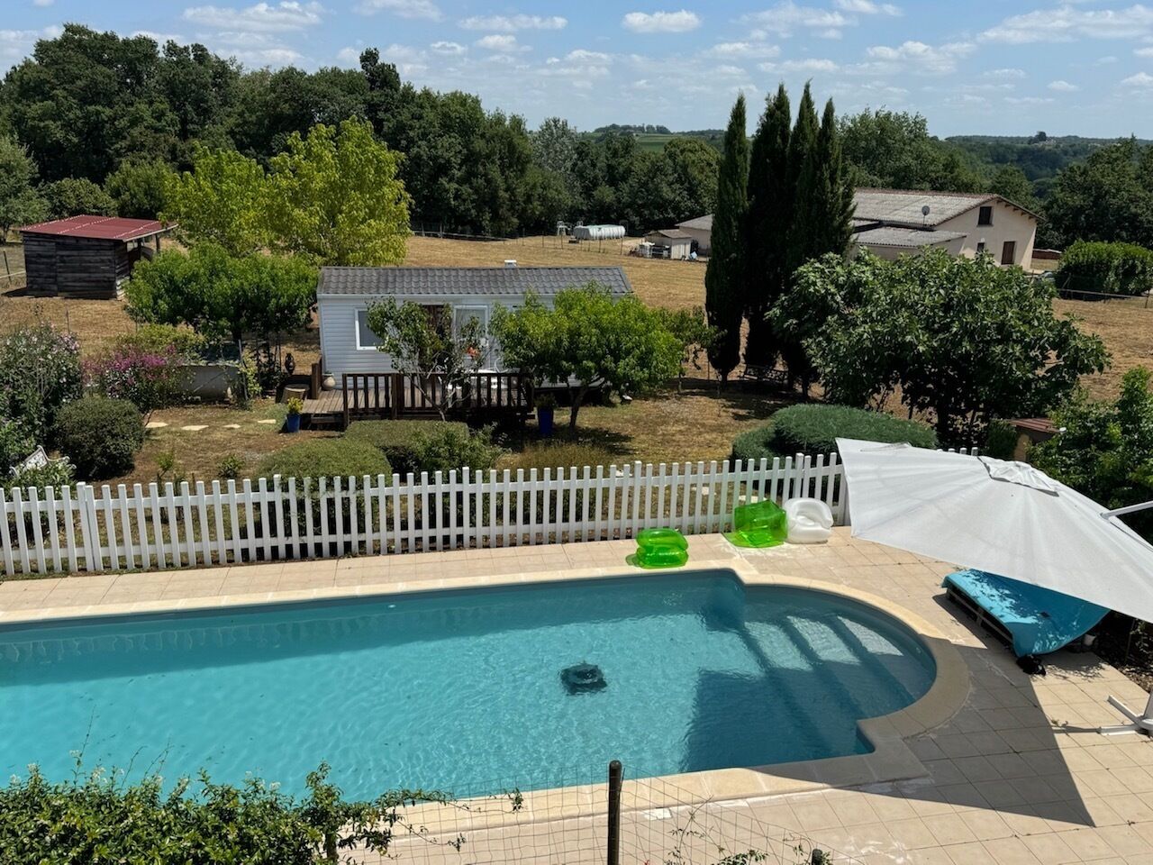 A swimming pool with a white fence, surrounded by trees and a small building.