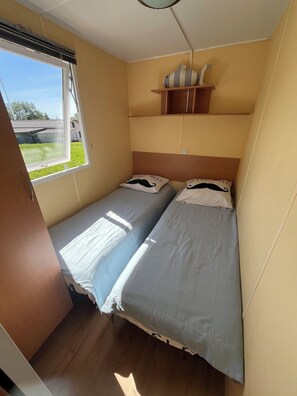 A compact room with two beds, a window, and a small shelf with decorative items. - Mobile Home in the Countryside (Juillac)