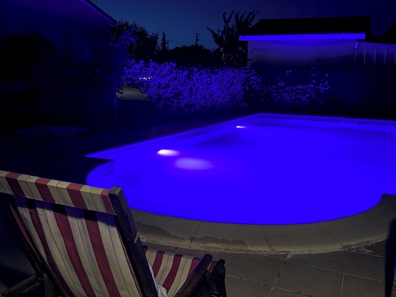 A swimming pool with a blue light feature, surrounded by a patio area with a chair.