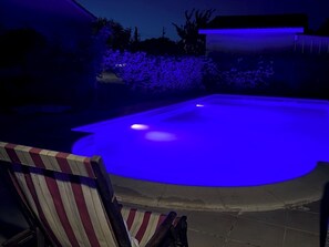 A swimming pool with a blue light feature, surrounded by a patio area with a chair. - Mobile Home in the Countryside (Juillac)