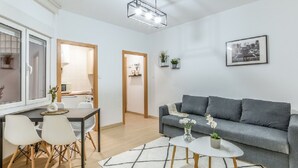 Moderni olohuone, jossa on harmaa sohva, ruokapöytä tuoleineen, avoimesta ovesta näkyvä keittiö ja seinällä oleva kehystetty taulu.