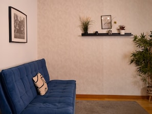 Un soggiorno con un divano blu, un quadro incorniciato appeso al muro, uno scaffale con oggetti decorativi e una pianta grande.