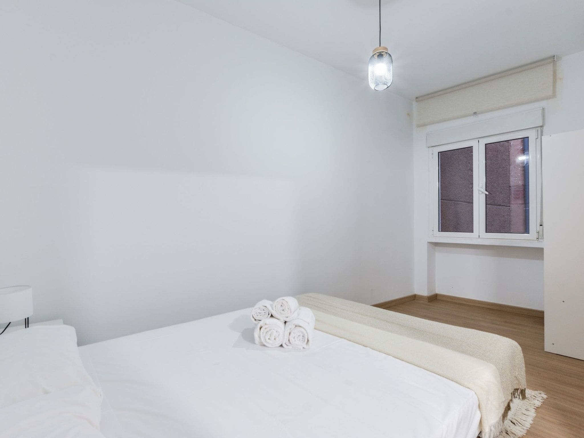 Un dormitorio minimalista con una cama, un ventanal con persianas y una lámpara colgante.