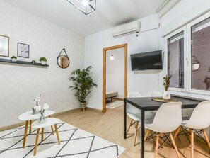 A modern living room with a dining area, a TV mounted on the wall, and a large window.