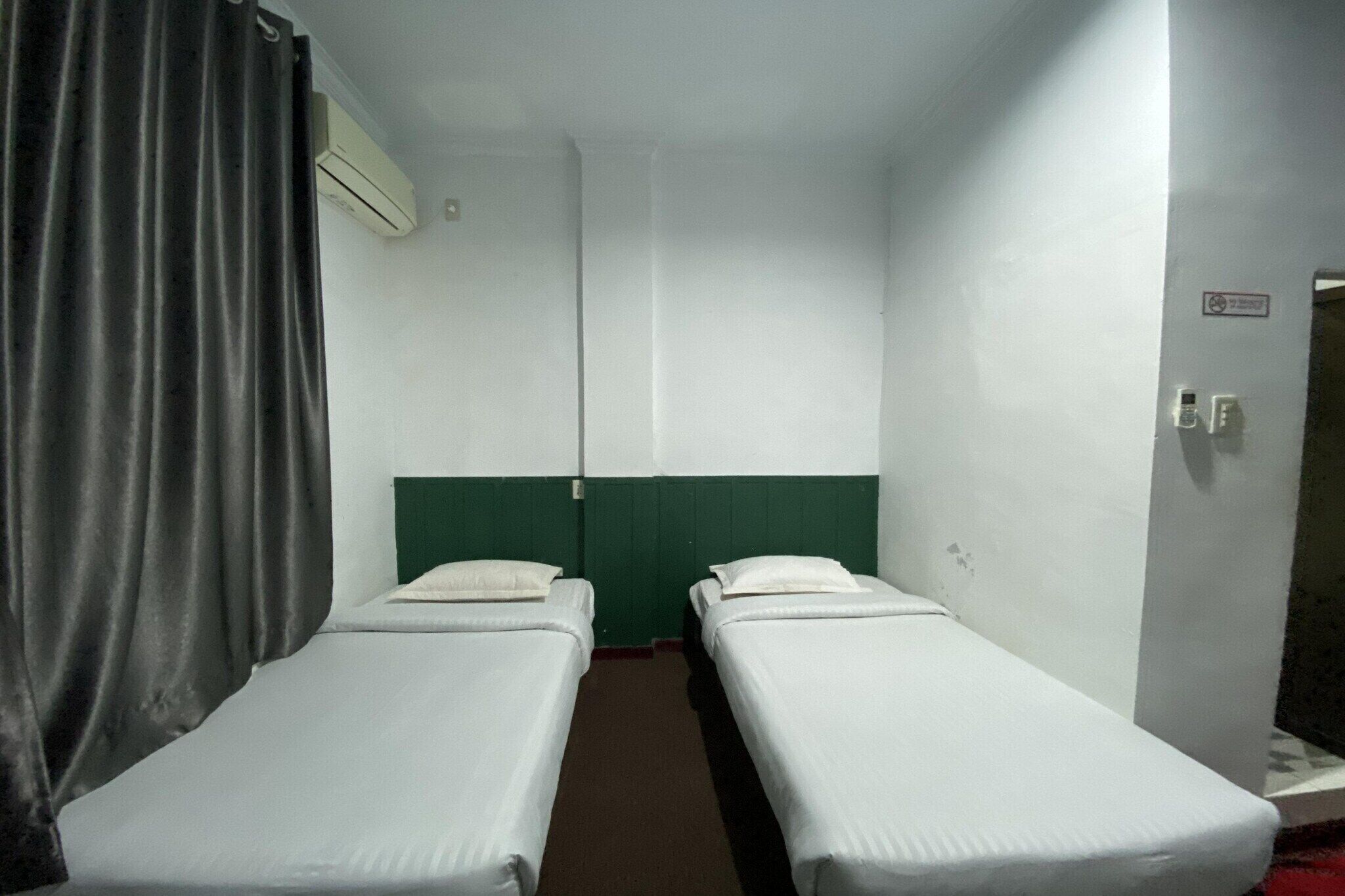 Ein kleines Hotelzimmer mit einem Bett für eine Person, zwei Einzelbetten, einem Vorhang, einer Klimaanlage und einer Tür mit Lichtschalter.