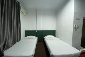 A small, single-room hotel room with two single beds, a curtain, an air conditioning unit, and a door with a light switch. - Quanza Hotel • Heritage Family Suite (Banda Aceh)