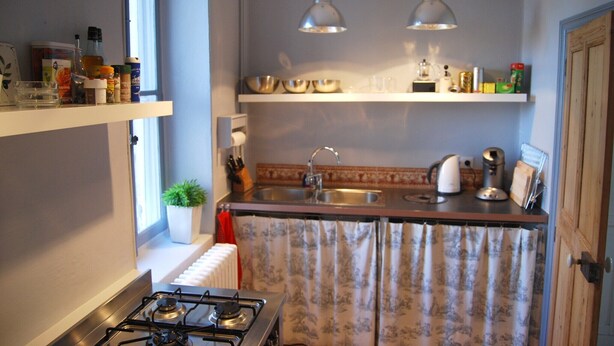 Een keuken met een gasfornuis, twee hanglampen, een spoelbak en verschillende keukengadgeten op planken.
