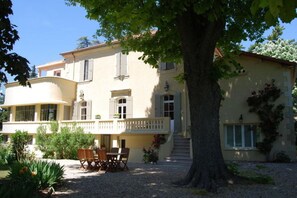 Ein zweistöckiges Gebäude mit Balkon, ein großer Baum davor und Sitzgelegenheiten im Freien.